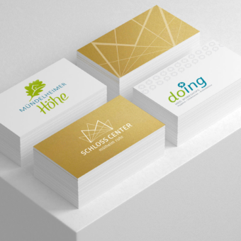 Drei Stapel Visitenkarten mit modernem Corporate Design liegen nebeneinander auf einer weißen Oberfläche. Die Karten zeigen verschiedene Logogestaltungen: links das Logo „Mündelheimer Höhe“ in Grün und Blau mit stilisiertem Baum, mittig das Logo „Schloss Center Mülheim Ruhr“ in Weiß auf goldfarbenem Hintergrund und rechts das Logo „doing Med. Workshops“ in Blau und Grün mit medizinischem Kreuz-Symbol.