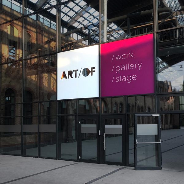 Foto des gläsernen Eingangsbereichs des Alten Schlachthofs in Offenbach, das große Logo der Art/Of Gallery ist über dem Eingang auf die Fensterfächen foliert.